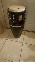 Full conga drum standing on tile floor showing black lacquered body with tuning hardware and natural skin head.