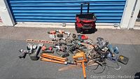 Overview image of all items in the lot arranged outside in daylight on asphalt with a blue industrial door background. Includes power tools, cords, tool bag, and accessories.