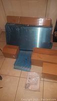 Full view of the new stainless steel under cabinet range hood wrapped in blue protective film, along with several cardboard boxes containing accessories and parts.