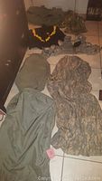 Five military gear pieces including 2 jackets, 1 vest, sleeping bag cover, and green bag on tiled floor