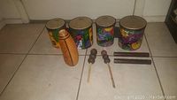 Overview photo showing two sets of Remo kids congos decorated with jungle and animal images, a Jam Town wooden gourd guiro, vintage wooden shakers on sticks, and Jam Town wooden sticks.