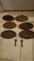Six oval WKM cast iron sizzle dishes arranged in two rows of three with ridged grilling surfaces, paired with six wooden oval bases showing wear and scratches, and two metal carry handles at bottom center.