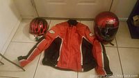 Red and black pilot motorcycle jacket laid flat with three helmets arranged around it: one red HJC helmet, one smaller black helmet with flame decals, and one smaller red and black helmet.