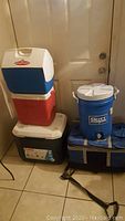 Stack of four hard coolers including a blue Playmate cooler on top, red and white Coleman cooler, large dark gray and white Coleman cooler on bottom; beside them a blue Gott water cooler with white lid on a soft cooler bag.