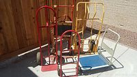 Lot showing three hand trucks and two platform carts lined up on concrete outside next to wooden fence; visible wear and scuff marks on metal and paint.