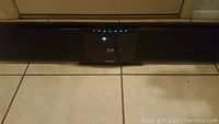 Front view of Samsung soundbar with Blu-ray player visible on front center panel, sitting on tiled floor.