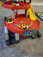 Front and side view of a red multistory toy car parking garage with multiple small toy cars on top and around it.