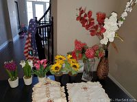Photo of assorted artificial flowers including sunflowers, tulips, and other blooms in small pots along with embroidered table linens.