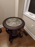 Round metallic wood-coloured end table with decorative metal scrollwork under glass top, placed on wooden floor next to window.