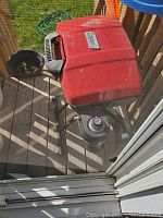 Top-down view of the Coleman Sports foldable grill with red lid, black body, and propane tank underneath, placed on wooden deck.