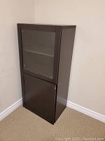 View of one black cupboard with glass door on top and solid door on bottom, closed and placed against a beige wall corner