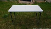 White foldable plastic table set up outdoors on grass, showing table top and metal legs.