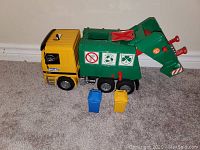 Photo of the toy garbage truck with yellow cab and green waste compartment positioned on hard floor, with two small trash cans in front, one blue one yellow.