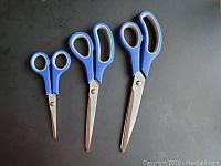 Three pairs of scissors in small, medium, and large sizes with blue and black handles on a black surface