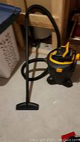 Shop-Vac vacuum cleaner with hose and nozzle attached, black and yellow in color, shown on a concrete floor next to a laundry hamper and shelving.