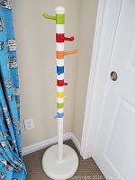 Photo of a white standing clothes hanger with multiple rainbow color hooks on a segmented pole and round base against a wall near a blue curtain.