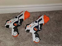 Two white and orange Nerf guns with black handles on carpeted floor