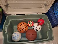 Five sports balls (basketballs and soccer balls) in a plastic storage bin. Visible brands include Spalding on the orange basketball.