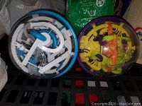 Photo of two Perplexus 3D maze puzzle spheres, one blue/white and one purple/yellow, placed side by side on a black grid surface.