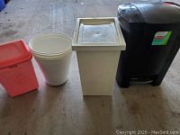 Four plastic containers lined up including pink floral trash can, white bucket, white container with lid, black trash can with foot pedal