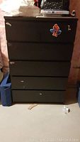 Front view of black IKEA MALM dresser showing 5 drawers with minor surface wear and stickers, against a plain background.