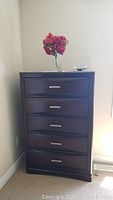 Front view of the dark brown chest of drawers with five drawers and silver handles, topped with a vase of red flowers and a decorative item.