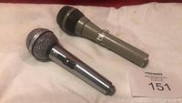 Two handheld microphones side by side on a white surface, one metallic chrome with mesh grille, the other matte gray with black foam windscreen.