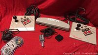 Two Nintendo NES Advantage joystick controllers, one Retro USB 12 MultiTap adapter, a Nintendo NES controller, a Nintendo NES controller connector adapter and a power adapter on red background.