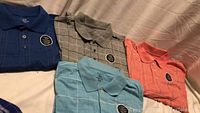 Four men's polo shirts laid out to show front and pattern. Colors: navy blue, grey, light blue, coral. Each shirt has a chest pocket on left side.