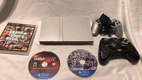 Photo of Sony PS2 Slim console, silver controller with cable, black controller, and Grand Theft Auto V PS4 game case.