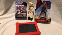 Three toys and one tablet displayed on flat surface: Spider-Man action figure, Spider-Man Blast Go motorcycle toy, sailor plush doll, and tablet.