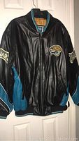 Full view of the front of the black leather Jacksonville Jaguars jacket showing teal accents, ribbed collar and embroidered jaguar logo on left chest.