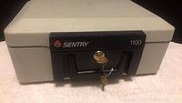 Closed view of the Sentry 1100 safe showing the locked front panel with key inserted in the lock and the black handle.