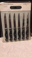 Photo of six Mainstays steak knives with black handles and serrated stainless steel blades in original packaging.