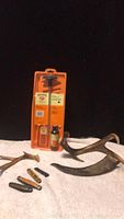 Full view of all items including the Hoppes pistol cleaning kit, four pocket knives arranged in front, and two pieces of deer antlers