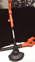 Black & Decker string trimmer showing orange handle, metal extendable shaft, and trimming head with guard.