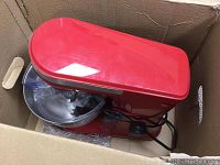 Red Amazon Basics stand mixer inside a cardboard box, showing stainless steel bowl, splash guard, and power cord. Slight scuffs visible.
