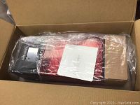 Opened cardboard box showing printer wrapped in protective plastic, red cover and black base visible