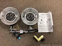 Full view of two circular radiant heating elements with protective guards attached to a horizontal metal pipe, control knob, flexible hose, bracket, and instruction manual on carpet.