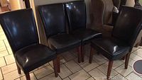 Photo showing four black faux leather dining chairs with wooden legs arranged together on a tiled floor.