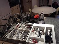 Overview of the kitchen items including pots, pans, white utensil holders, and cutlery trays with various forks, spoons, knives, and kitchen tools.