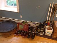 Wide shot of lot showing mini trampoline, bocce balls and case, child bike seat, two pairs of hockey skates, dart board, broom, and white bucket.