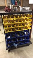 Front view of metal tool cart with several rows of yellow plastic bins filled with assorted hardware and blue bins at the bottom with larger components. A red mechanical part and plastic containers are on top shelf.