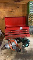 Red metal tool box with drawers and two electric drills placed in front of it.