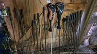 Wide view of multiple left handed golf clubs including irons, drivers, and hickory shaft clubs leaning against wooden wall.