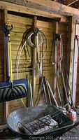 Various garden tools hanging on wood wall including shovels, rake, hose, hedge clippers and wheelbarrow in foreground