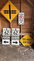 Seven road signs arranged against a wooden wall, including railroad crossing, Do Not Enter, Highway 62 directional signs, and fire route sign.