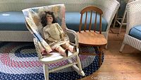 Full view of the two child-sized chairs with the antique doll sitting on the white wicker rocking chair, quilt visible behind.