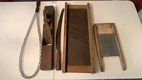 All five antique farm tools arranged on a table: wooden wood plane, long knife, wooden rectangular tool with diagonal metal braces, wooden framed wash board, and woven wooden carpet beater.