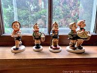Full view of all four Hummel figurines lined up on a wooden surface by a window, showing the Boy with basket, Boy with backpack, Boy with walking stick, and Boy and girl figurines.